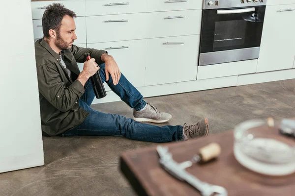 Foco seletivo de homem triste com garrafa de vinho sentado no chão da cozinha — Fotografia de Stock