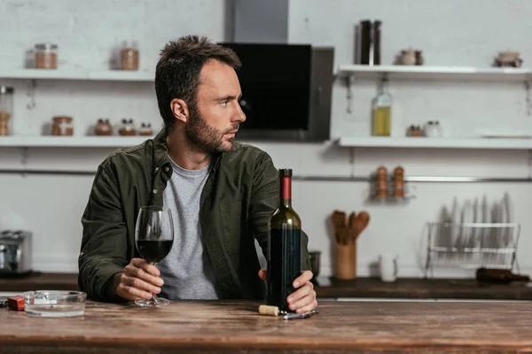 Uomo con bicchiere di vino e bottiglia guardando lontano al tavolo della cucina — Foto stock