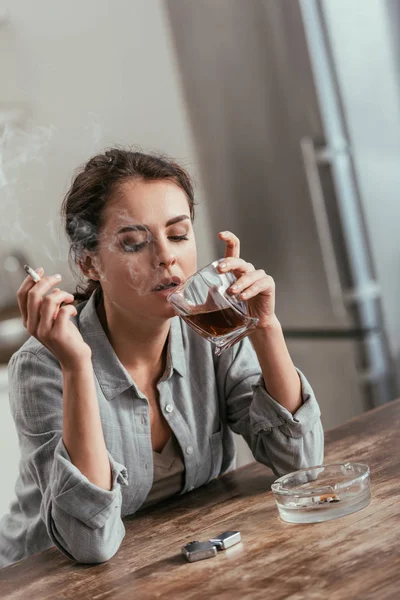 Mujer fumando cigarrillo y sosteniendo vaso de whisky en la mesa - foto de stock