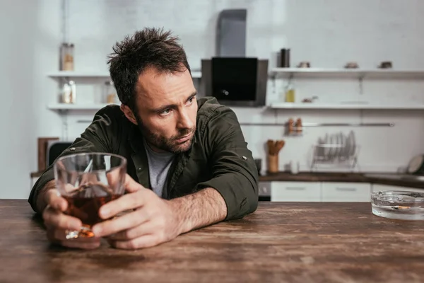 Focus selettivo dell'uomo preoccupato che tiene il bicchiere di whisky accanto al posacenere sul tavolo della cucina — Foto stock