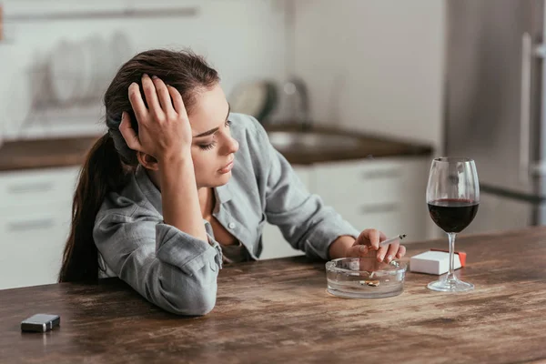 Femme inquiète fumant la cigarette à côté du verre de vin sur la table — Photo de stock