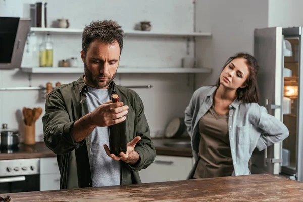 Selective focus of alcohol depended man with wine bottle and sad wife on kitchen — Stock Photo