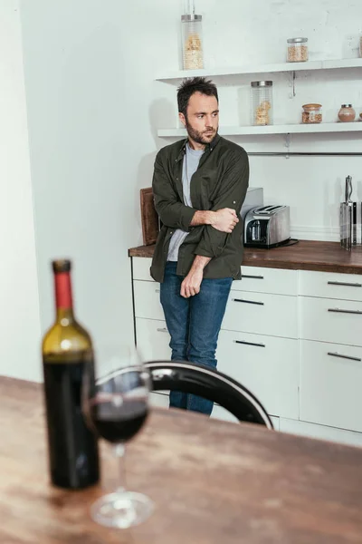 Selective focus of sad man standing on kitchen and wine glass with bottle on table — Stock Photo