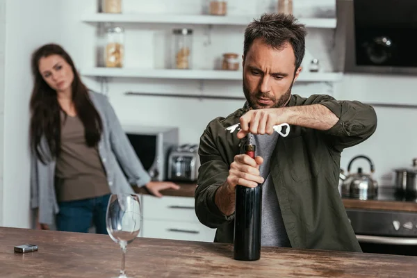 Selective focus of alcohol depended man opening wine bottle and worried wife on kitchen — Stock Photo