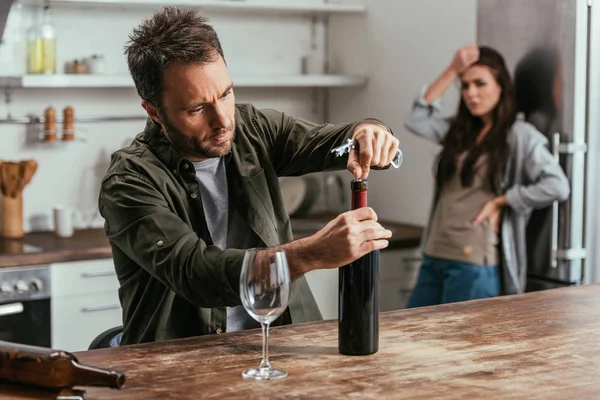 Selective focus of alcohol depended man opening wine bottle and upset wife on kitchen — Stock Photo