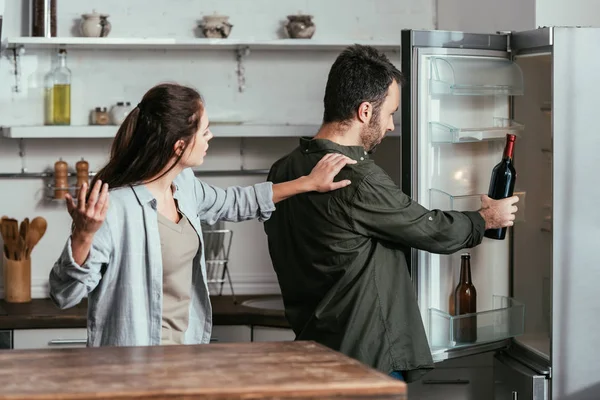 Angry woman quarreling with husband taking wine bottle from fridge — Stock Photo