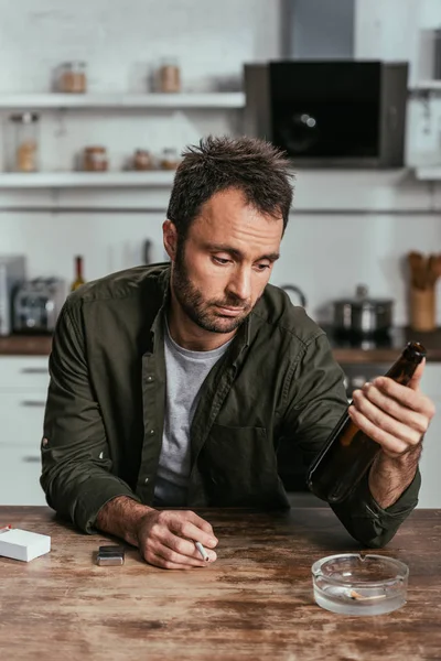 Uomo sconvolto con alcol dipendeva tenendo sigaretta e bottiglia di birra a tavola — Foto stock