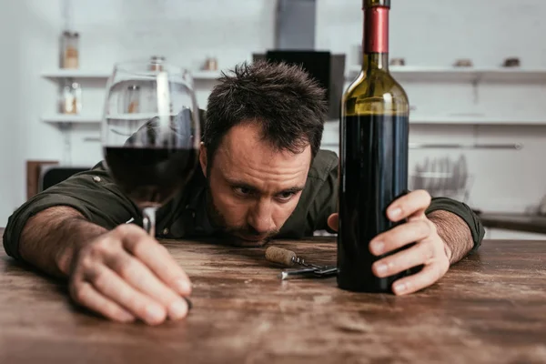 Foco seletivo de homem triste segurando copo de vinho e garrafa na mesa da cozinha — Fotografia de Stock