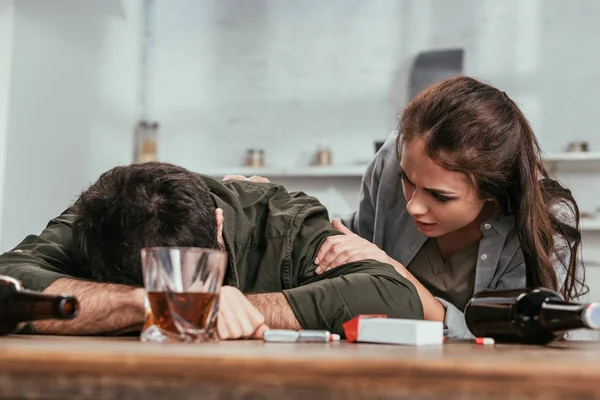 Enfoque selectivo de la mujer hablando con el marido borracho al lado de botellas y cigarrillos en la mesa - foto de stock