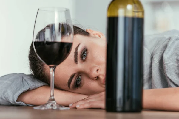 Foco seletivo da mulher olhando para a câmera com copo de vinho e garrafa na mesa — Fotografia de Stock