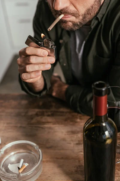 Cropped view of man lighting cigarette beside wine glass and bottle on table — Stock Photo