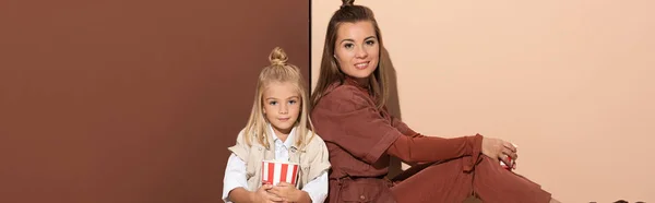 Panoramic shot of daughter with popcorn and smiling mother looking at camera on beige and brown background — Stock Photo