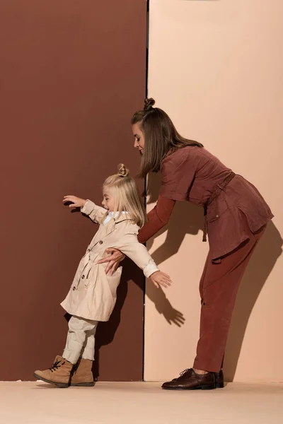 Seitenansicht einer lächelnden Mutter, die ihre süße Tochter auf beigem und braunem Hintergrund hält — Stockfoto