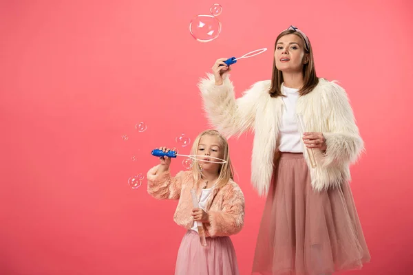 Daughter and smiling mother blowing soap bubbles on pink background — Stock Photo