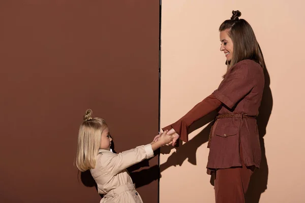 Seitenansicht von lächelnder Tochter und Mutter, die sich auf beigem und braunem Hintergrund an den Händen halten — Stockfoto