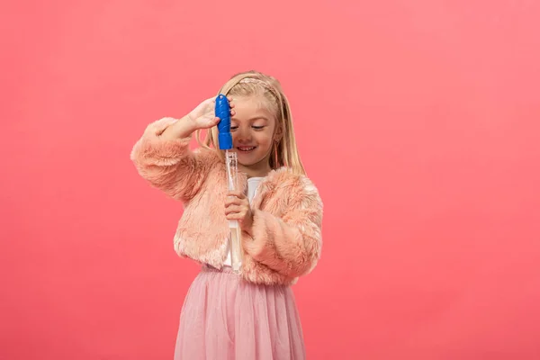 Lindo y sonriente niño sosteniendo botella con burbujas de jabón aislado en rosa - foto de stock