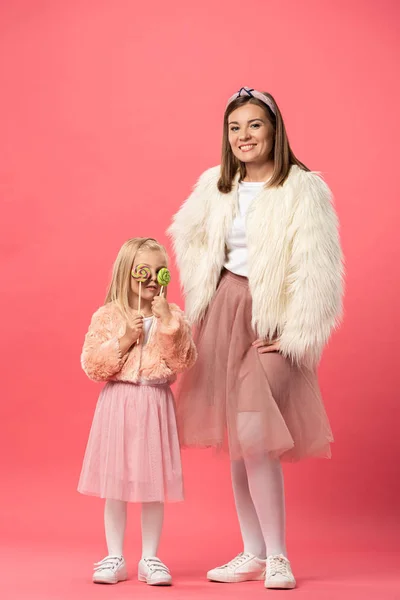 Daughter obscuring face with lollipops and smiling mother looking at camera on pink background — Stock Photo