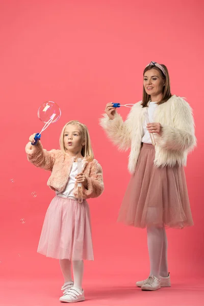 Hija y sonriente madre soplando burbujas de jabón sobre fondo rosa - foto de stock