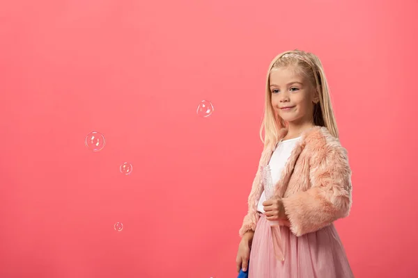 Милый и улыбающийся ребенок держит бутылку с мыльными пузырями изолированы на розовый — стоковое фото