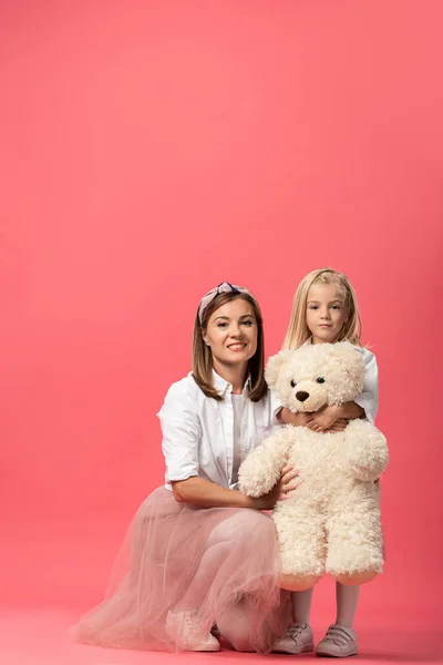Daughter with teddy bear and smiling mother looking at camera on pink background — Stock Photo