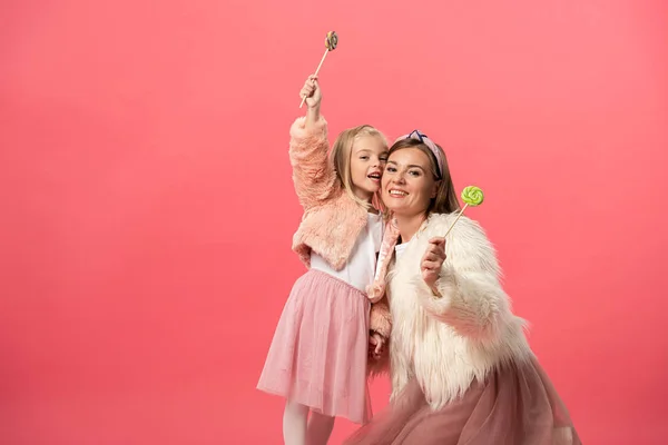 Sonriente hija y madre sosteniendo piruletas aisladas en rosa - foto de stock