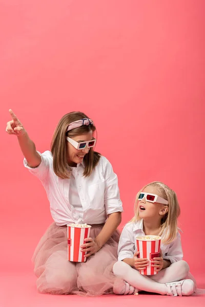 Conmocionada hija con palomitas de maíz y madre señalando con el dedo sobre fondo rosa - foto de stock