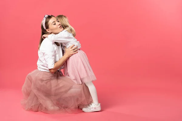 Hija abrazando atractiva y sonriente madre sobre fondo rosa - foto de stock