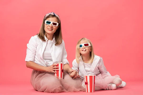 Hija comiendo palomitas de maíz y madre en gafas 3d mirando hacia otro lado sobre fondo rosa - foto de stock