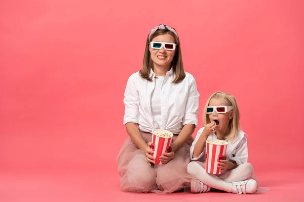 Hija comiendo palomitas de maíz y sonriente madre en gafas 3d sobre fondo rosa - foto de stock