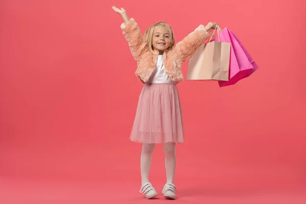 Lindo y sonriente niño con las manos extendidas sosteniendo bolsas de compras sobre fondo rosa - foto de stock