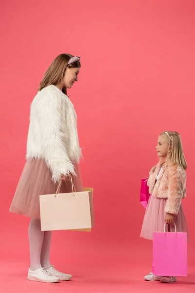 Vista lateral de sonriente hija y madre sosteniendo bolsas de compras sobre fondo rosa - foto de stock