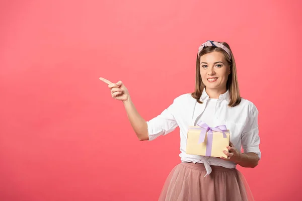 Attraktive und lächelnde Frau hält Geschenkbox und zeigt mit dem Finger isoliert auf rosa — Stockfoto