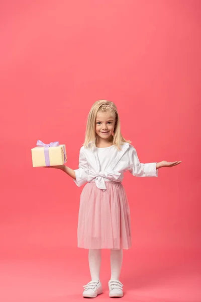 Niedliches und lächelndes Kind mit ausgestreckter Hand, die Geschenk auf rosa Hintergrund hält — Stockfoto