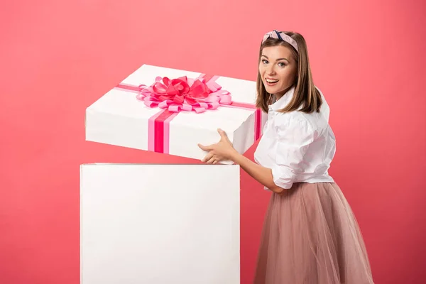 Attraktive und lächelnde Frau öffnet Geschenkbox isoliert auf rosa — Stockfoto