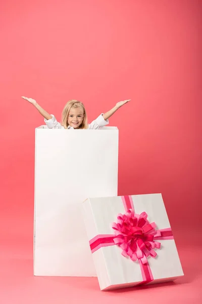 Милый и улыбающийся ребенок с протянутыми руками в подарочной коробке на розовом фоне — стоковое фото