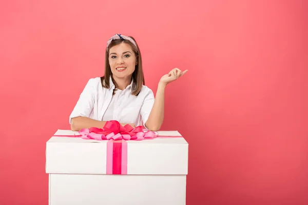 Attrayant et souriant femme debout près boîte cadeau et pointant avec doigt isolé sur rose — Photo de stock