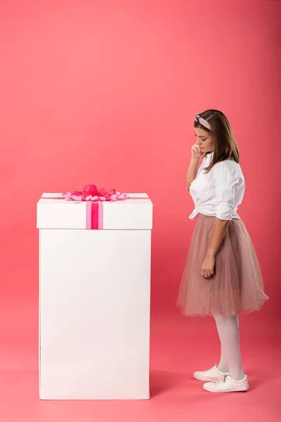 Mujer atractiva y pensativa de pie cerca de la caja de regalo sobre fondo rosa - foto de stock