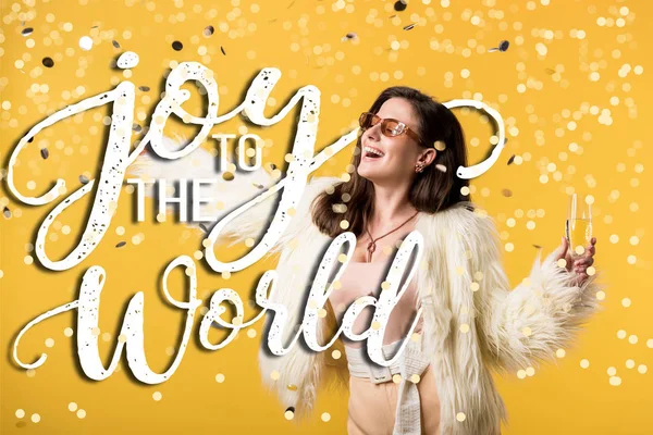 Excited party girl in faux fur jacket and sunglasses standing under confetti with champagne isolated on yellow with joy to the world illustration — Stock Photo