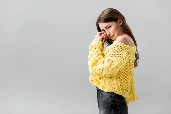 Chica molesta en suéter amarillo llorando, limpiando las lágrimas con la mano y mirando a la cámara aislada en gris - foto de stock