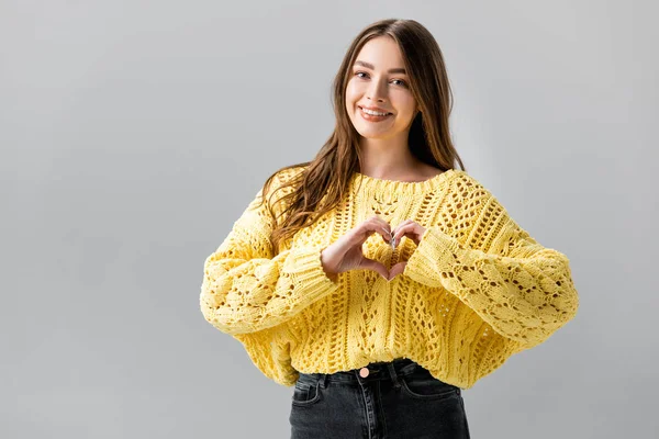 Sorridente ragazza in maglione giallo che mostra il segno del cuore isolato sul grigio — Foto stock