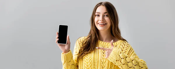 Панорамный снимок веселой молодой женщины, указывающей пальцем на смартфон с черным экраном, изолированным на сером — стоковое фото