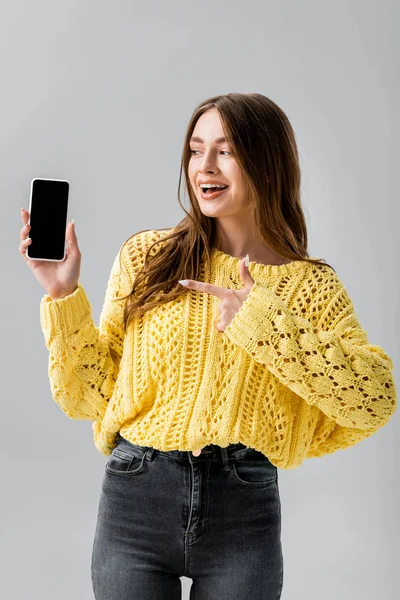 Bonita mujer en suéter amarillo sonriendo mientras señala con el dedo al teléfono inteligente con pantalla en blanco aislado en gris - foto de stock