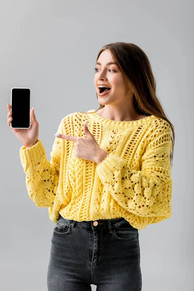 Fröhliche junge Frau zeigt mit dem Finger auf Smartphone mit leerem Bildschirm isoliert auf grau — Stockfoto