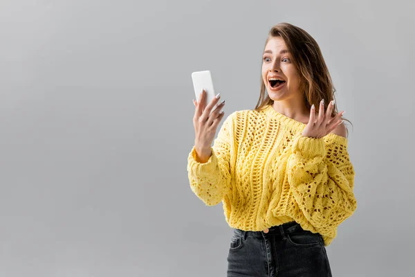 Chica sorprendida mostrando wow gesto durante el chat de vídeo en el teléfono inteligente aislado en gris - foto de stock