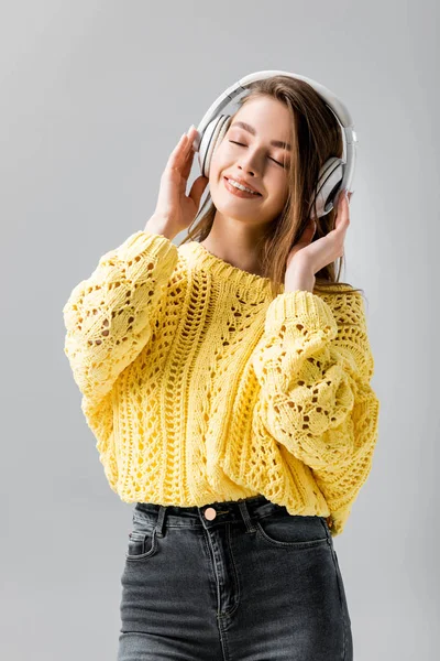 Chica atractiva y sonriente escuchando música en auriculares inalámbricos con los ojos cerrados aislados en gris - foto de stock