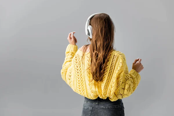 Visão traseira da menina em camisola amarela dançando enquanto ouve música em fones de ouvido sem fio isolados em cinza — Fotografia de Stock