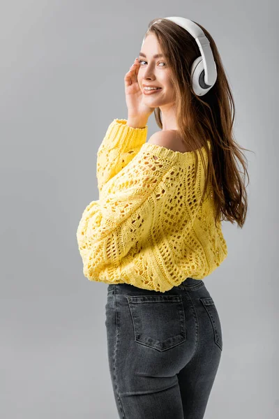 Menina atraente e sorridente olhando para a câmera enquanto ouve música em fones de ouvido sem fio isolados em cinza — Fotografia de Stock