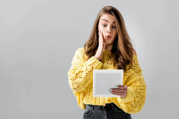 Chica sorprendida tocando la cara mientras mira tableta digital aislado en gris - foto de stock