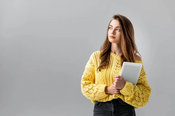 Mujer joven reflexiva mirando hacia otro lado mientras sostiene la tableta digital aislada en gris - foto de stock