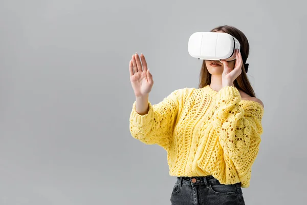 Mujer joven en suéter amarillo haciendo gestos mientras usa auriculares vr aislados en gris - foto de stock